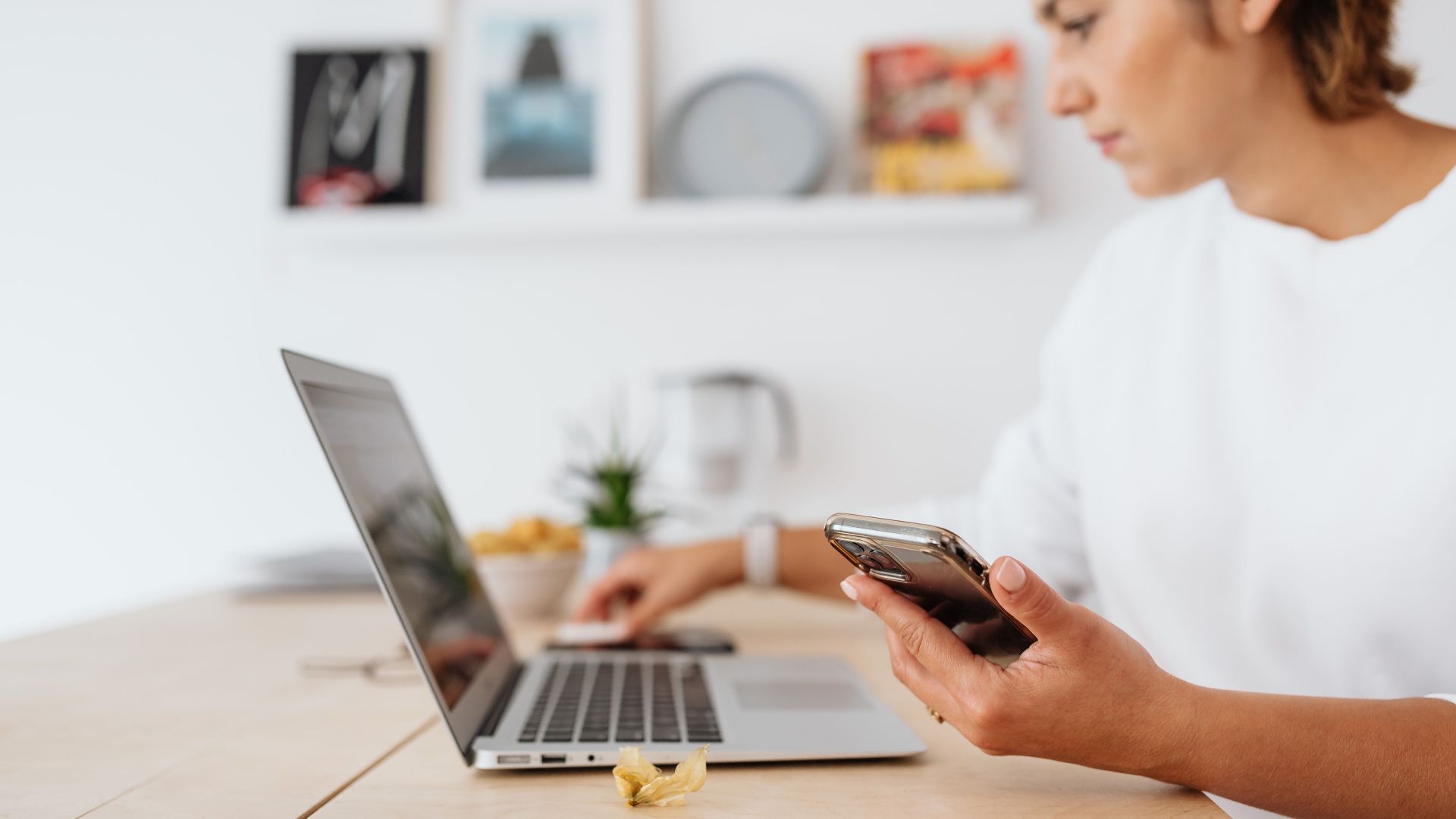 A woman in a white shirt using a laptop and smartphone at a wooden table.