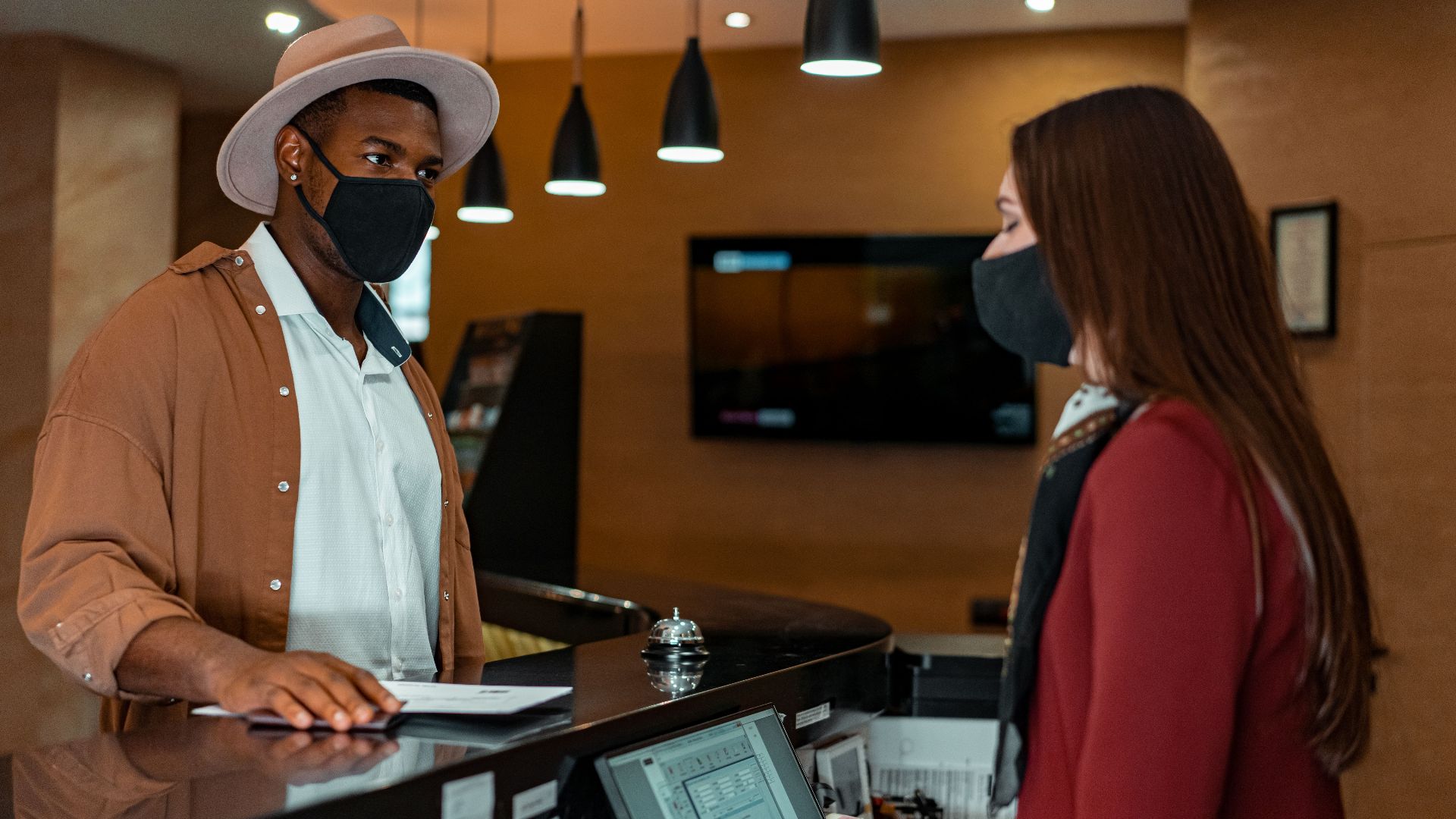 A man and woman in face masks checking in at a hotel reception, emphasizing safety protocols.