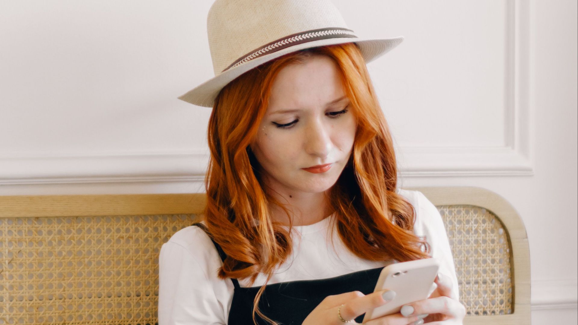 A young woman in a fedora hat texting on her smartphone while sitting with a laptop indoors.