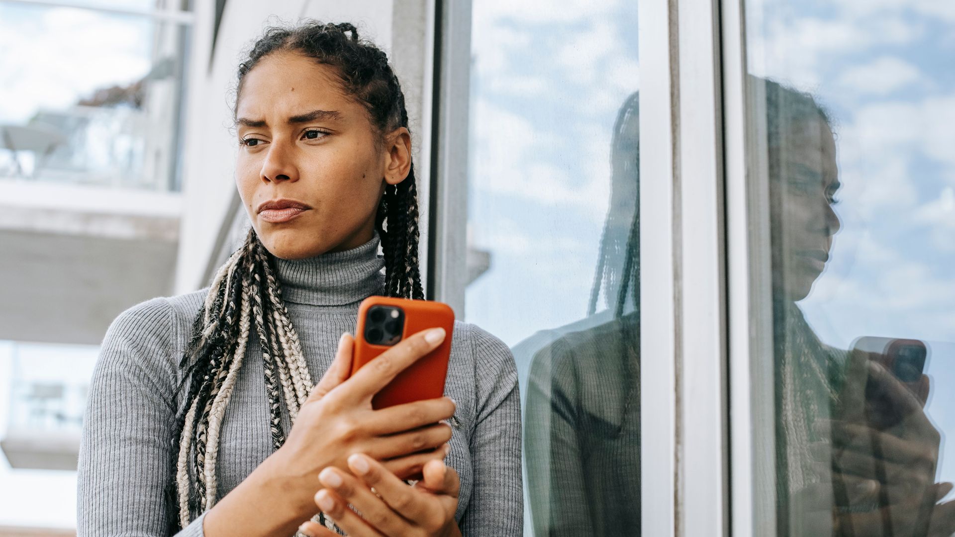 Low angle of concerned ethnic female with smartphone in stressful situation near glass wall