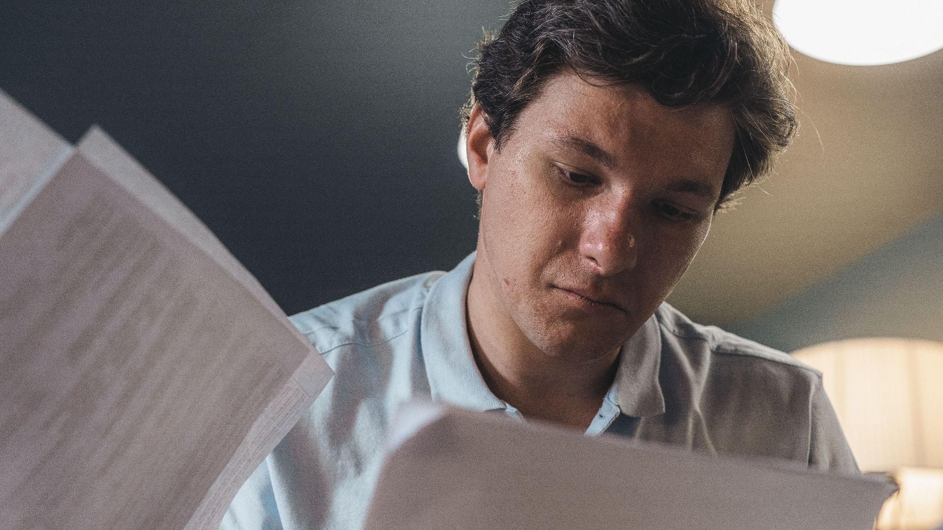 Serious man wearing a light blue shirt intently reading printed papers indoors under warm lighting.