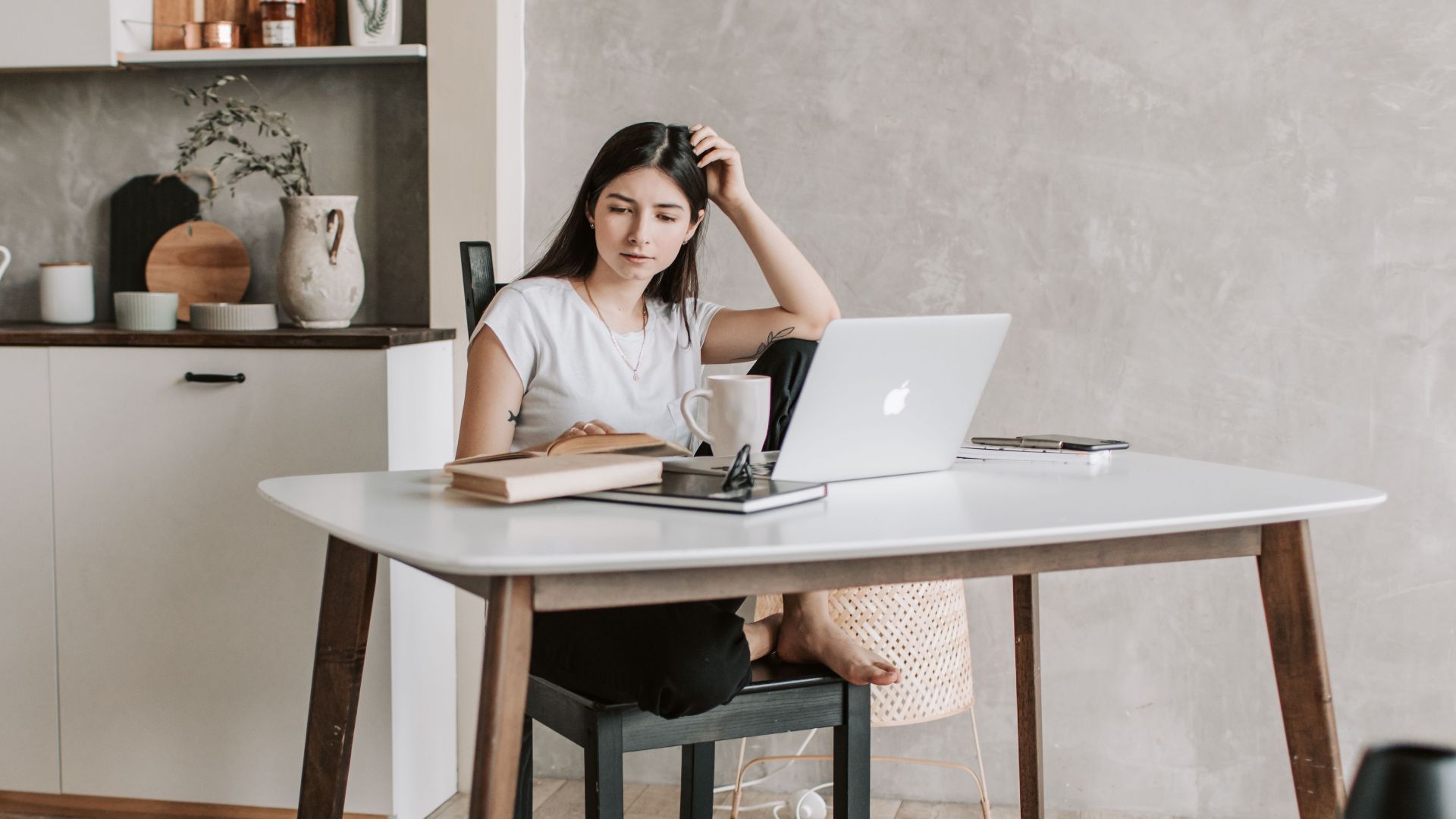 Concentrated female student sitting barefoot on chair at table with netbook and notebook while reading book during distance education in modern apartment with minimal interior in eco design