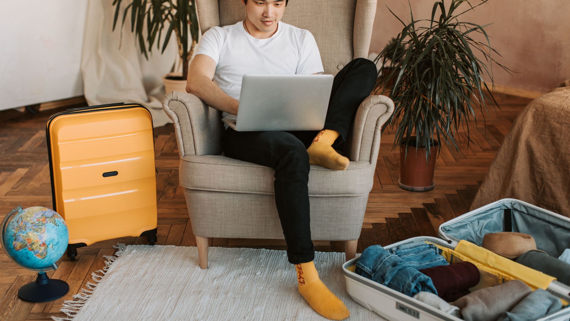 Man sitting on armchair planning travel, using laptop, with suitcases and globe nearby.
