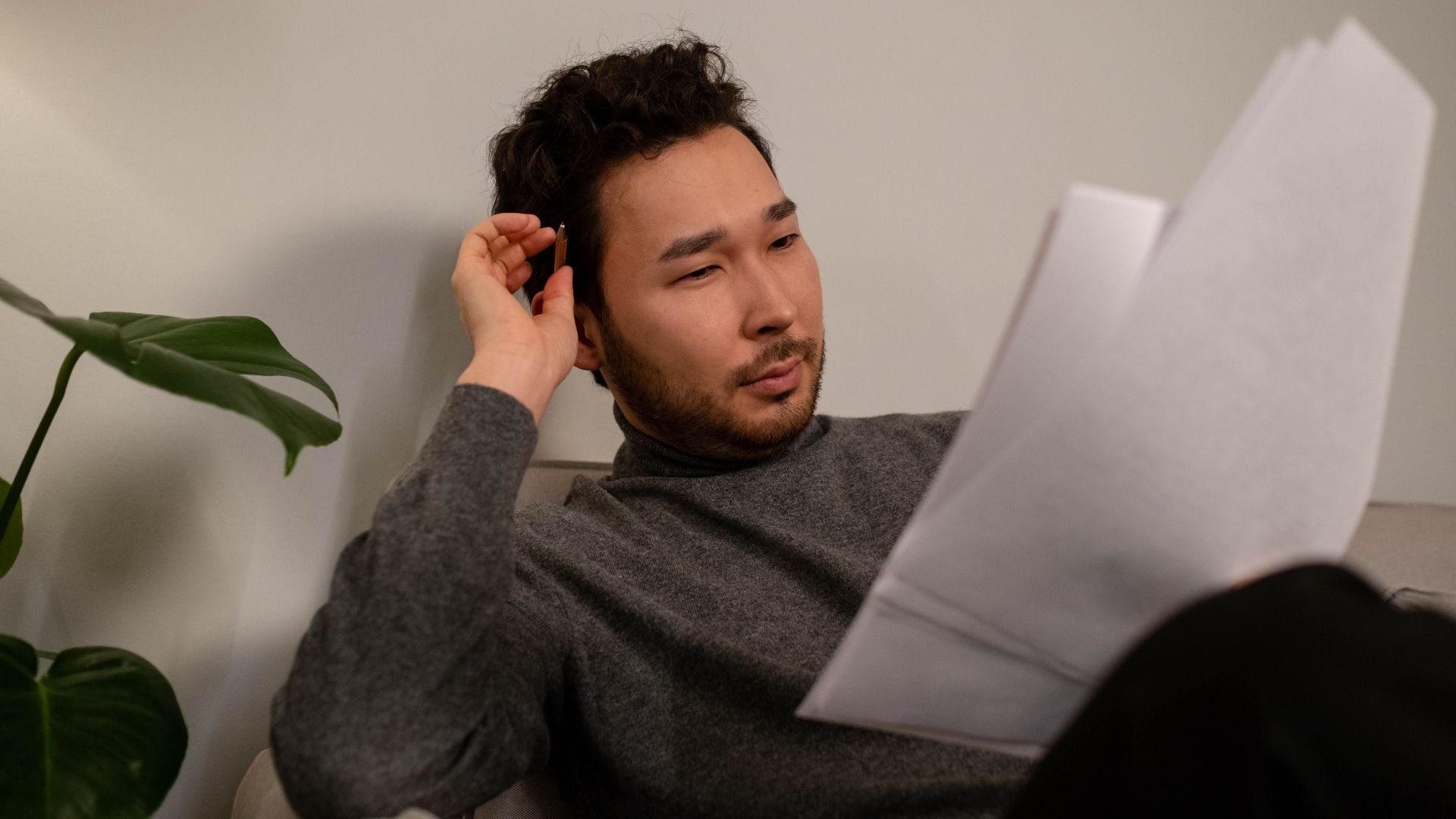 Asian man in a casual setting reading papers, sitting on a sofa with a plant nearby.