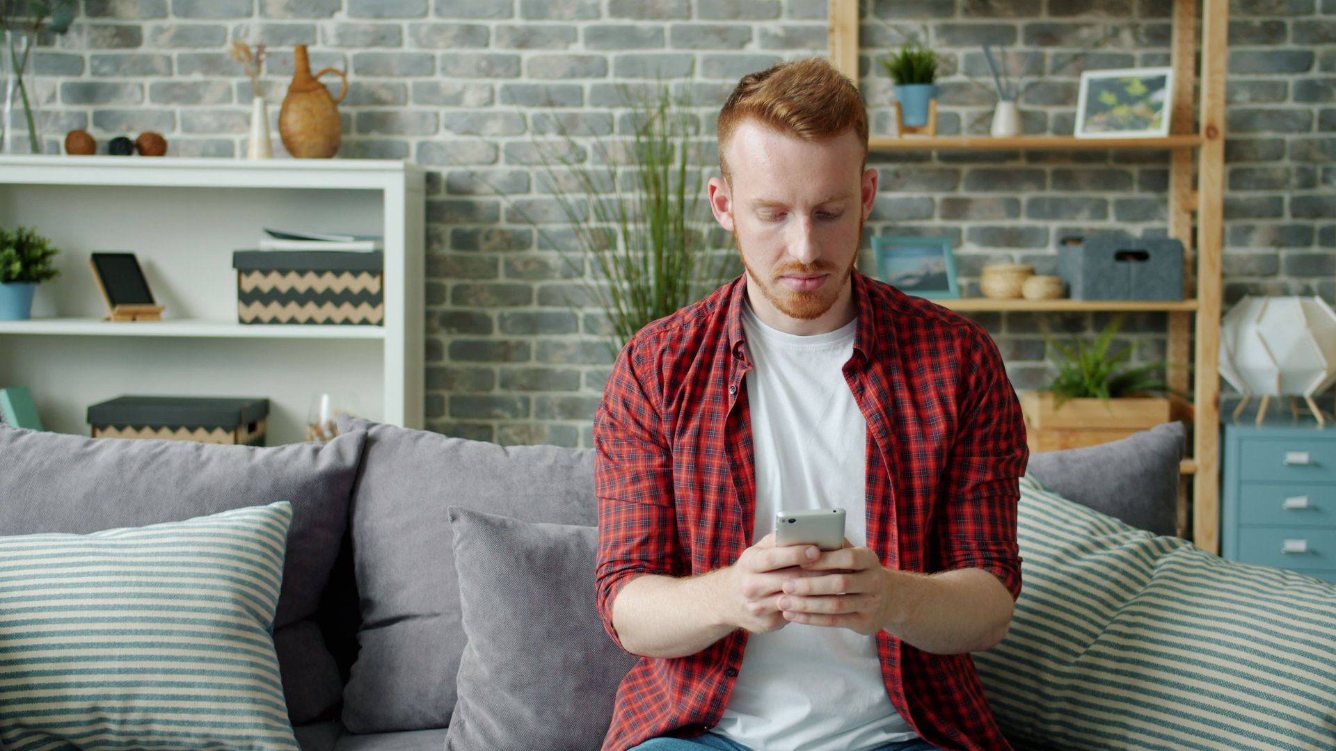 Young man in casual attire sitting on sofa using a smartphone indoors.