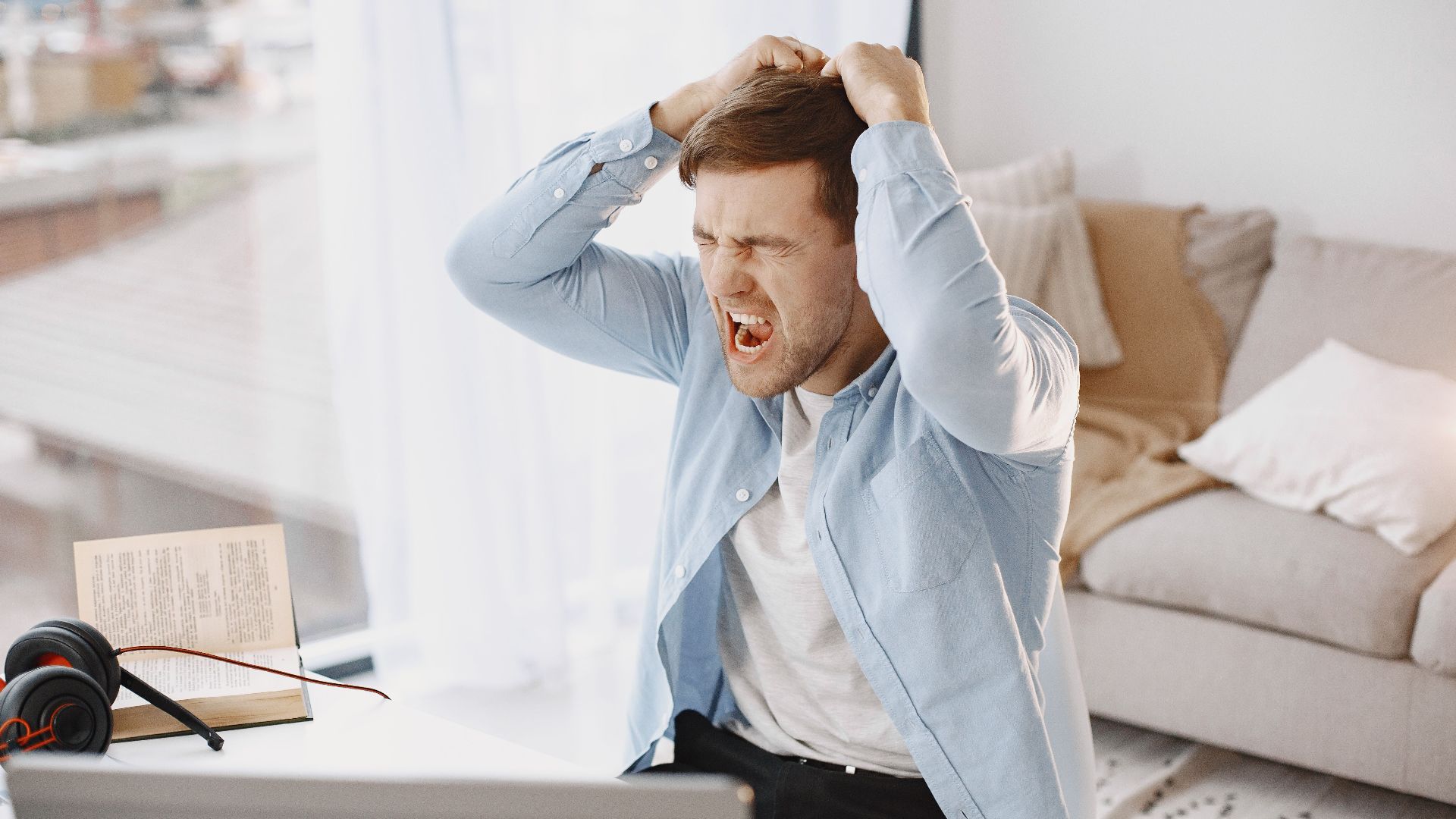 An upset man with mouth open, pulling hair at a desk with a laptop in a home setting.