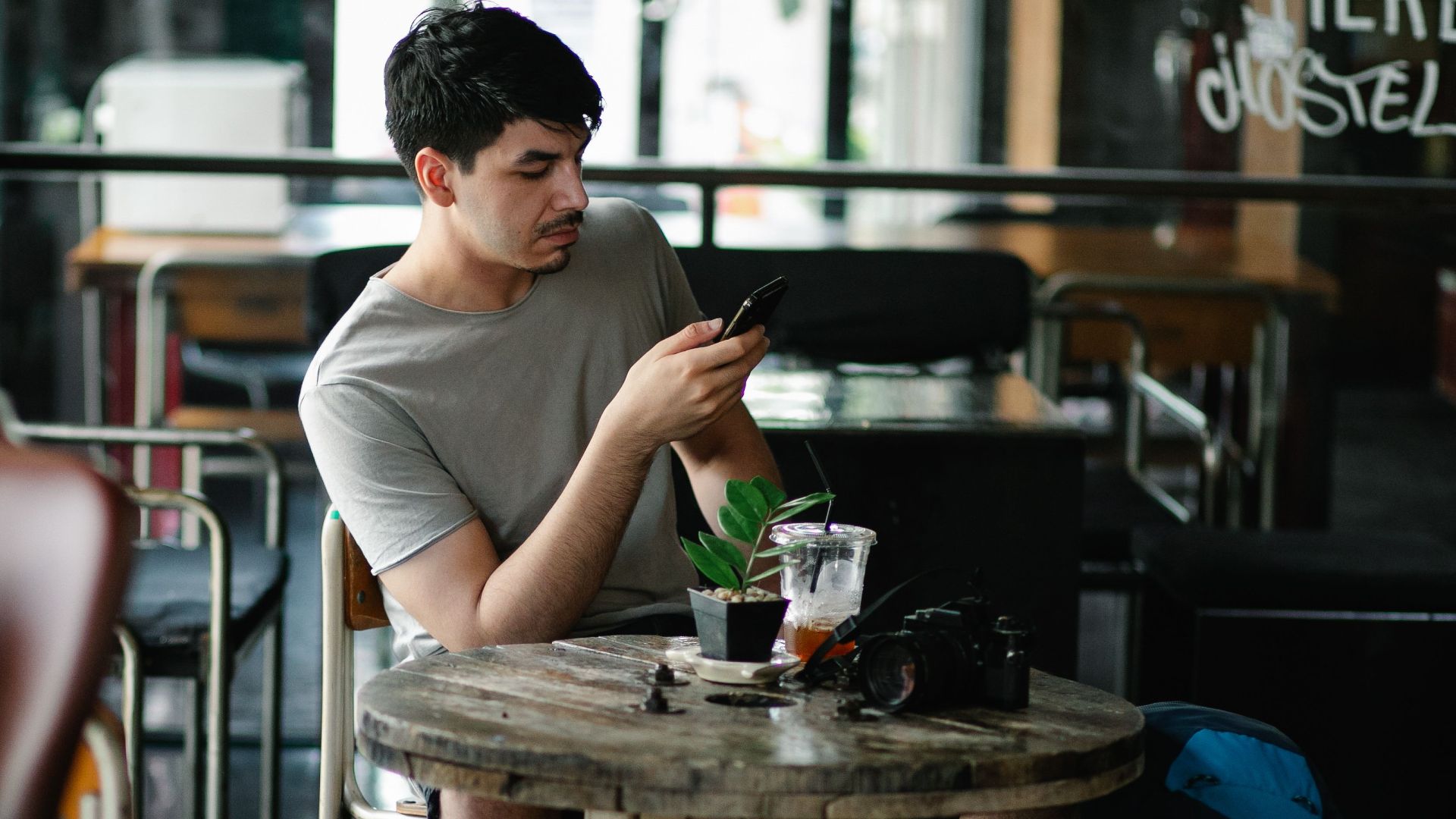 Concentrated male text messaging on cellphone while sitting at table with beverage and photo camera in cafeteria with blurred Asian woman