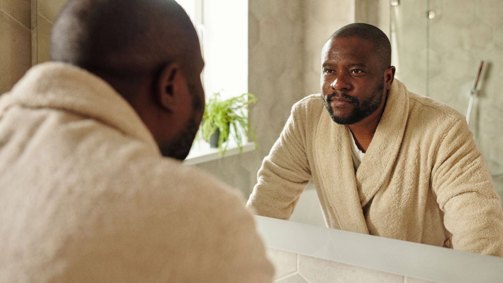 Adult man in a bathrobe looking at his reflection in a bathroom mirror. Morning routine concept.