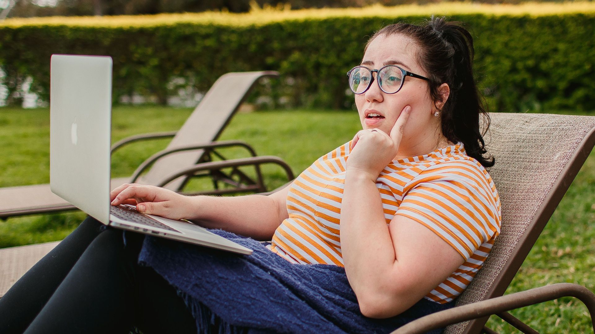 Woman relaxing on lounge chair with a laptop in a lush green garden, enjoying a sunny day.