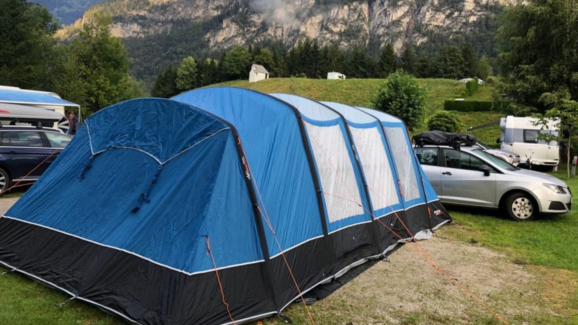 This picture shows a big blue tent at a campsite in the mountains. In the background, there are cars.