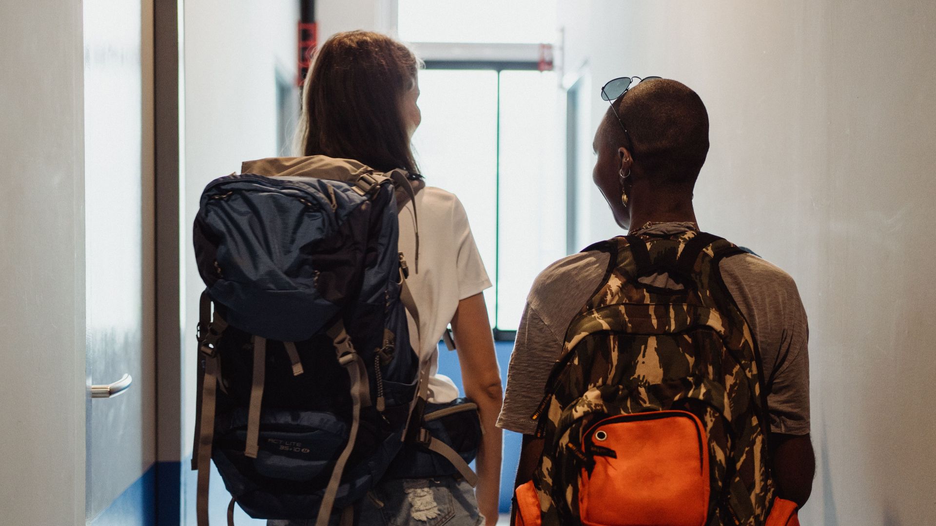Two friends walking in a hostel corridor with backpacks, embodying travel and adventure vibes.