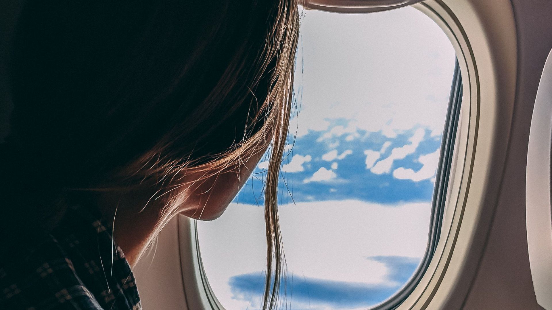 A woman looks out an airplane window, capturing a serene view of clouds and sky during a flight.