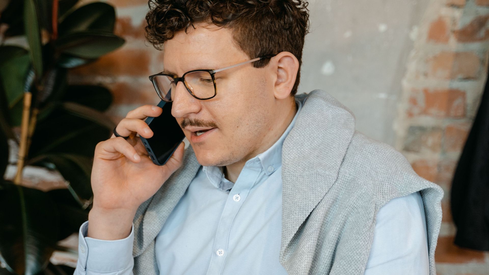 Man in casual attire talks on smartphone while working on laptop in a cozy workspace with brick wall.