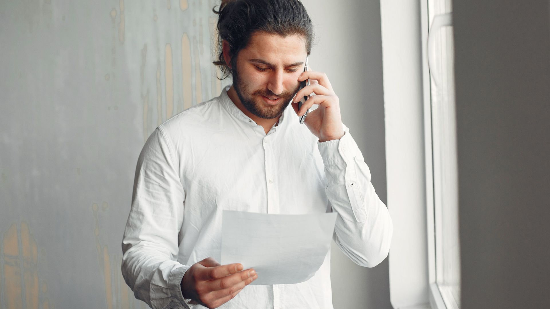 A young man in casual attire talks on the phone while holding a document indoors.