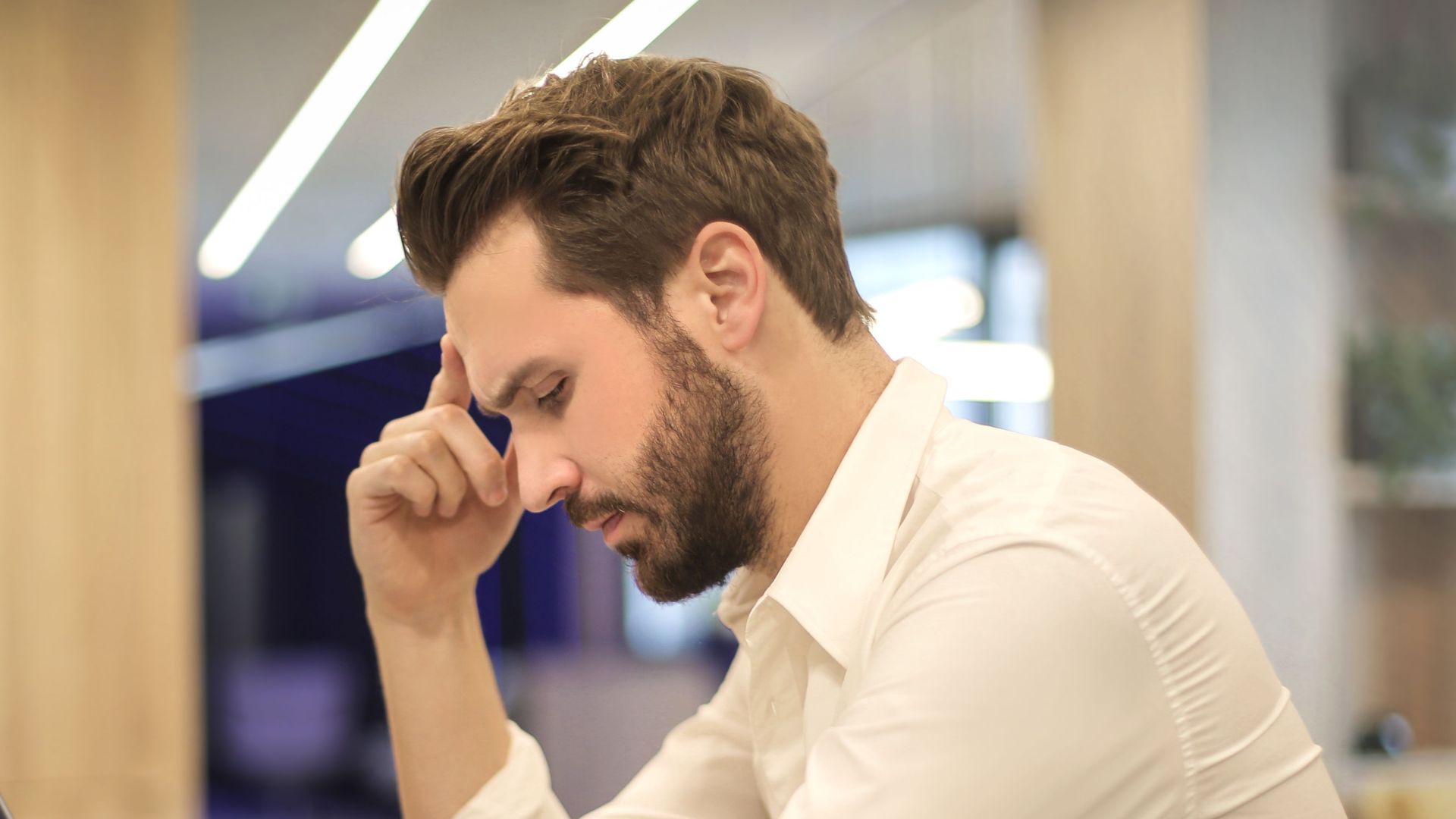 A thoughtful man in a shirt works on his laptop at a modern indoor office space.