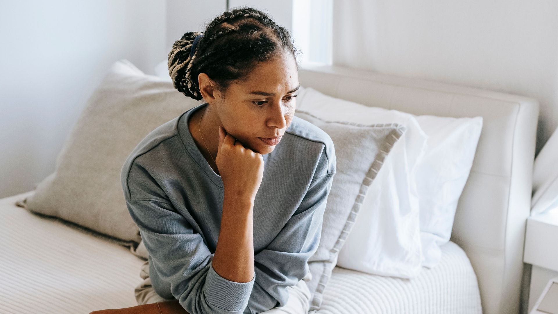 Serious young African American lady in casual clothes sitting on white bed and thinking while looking down in bedroom