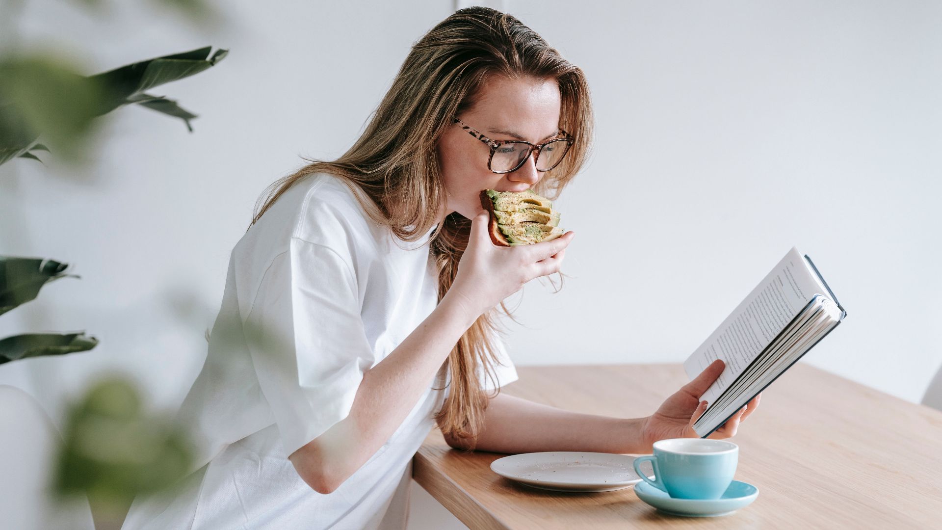 Side view of smart female in eyeglasses with long hair studying book at table with cup and plate