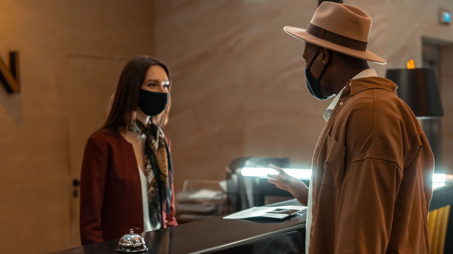 A man at a hotel reception talking to a staff member. Both wear face masks.