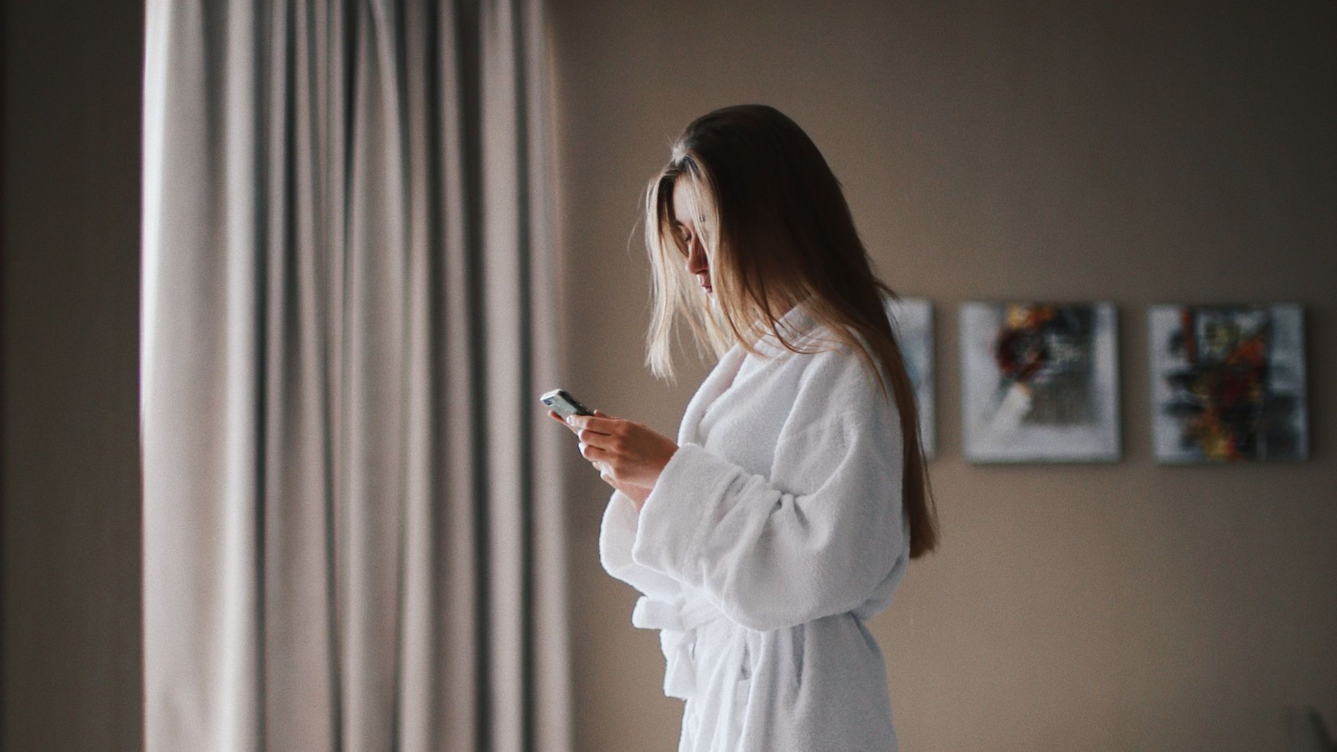 Woman standing in a hotel room wearing a bathrobe, focused on her smartphone for communication or relaxation.