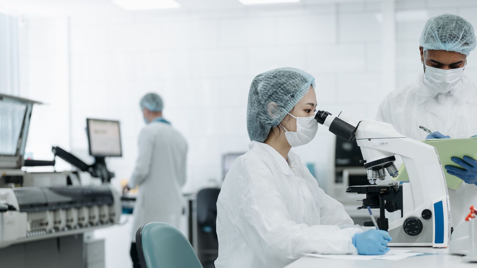 Scientists wearing protective gear conducting research in a lab, using a microscope to analyze samples.