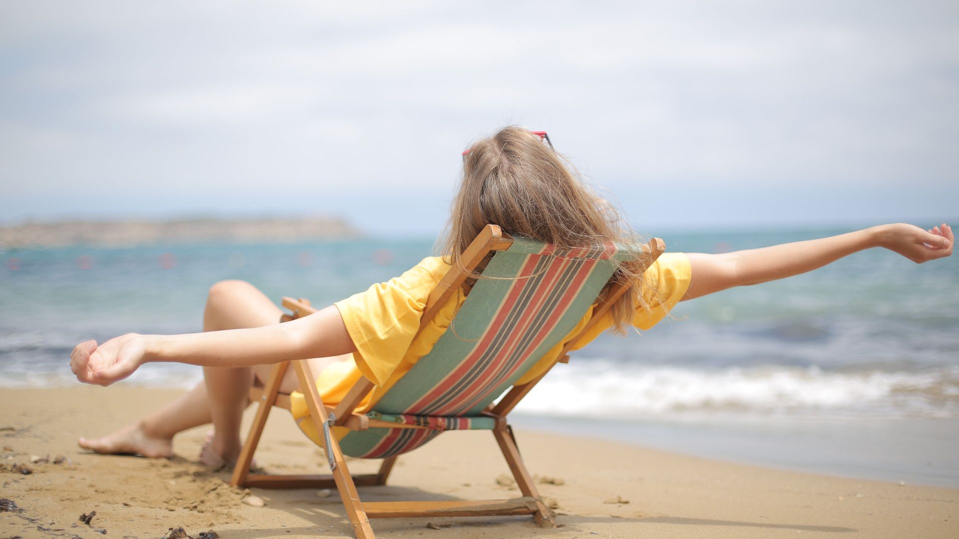 A woman in a yellow top relaxes on a folding chair at a sandy beach near the shore.