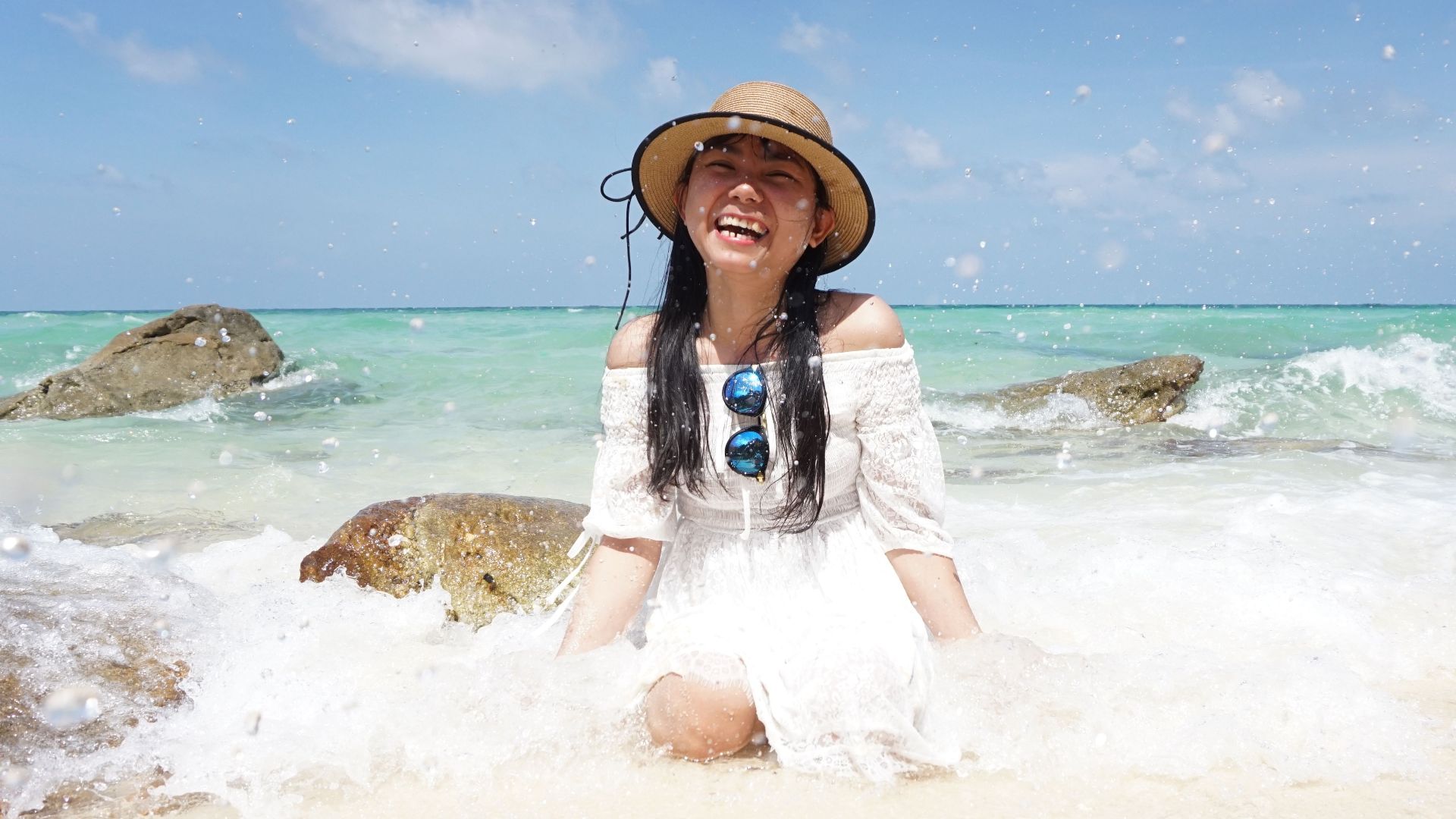 Asian woman smiling joyfully while seated on a sunny tropical beach with splashing waves.