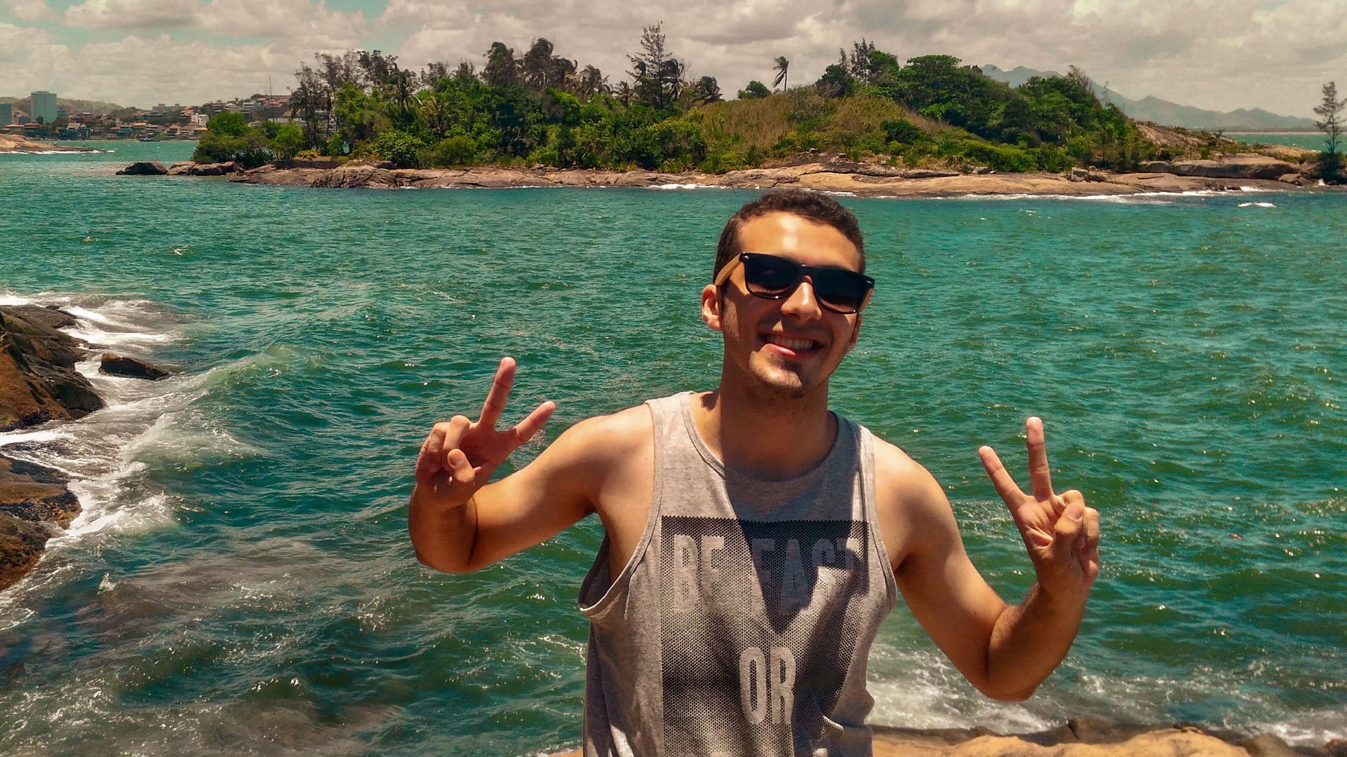 A man in sunglasses enjoys a sunny day at Praia do Canto, Brazil, showing peace signs on a rocky shore.