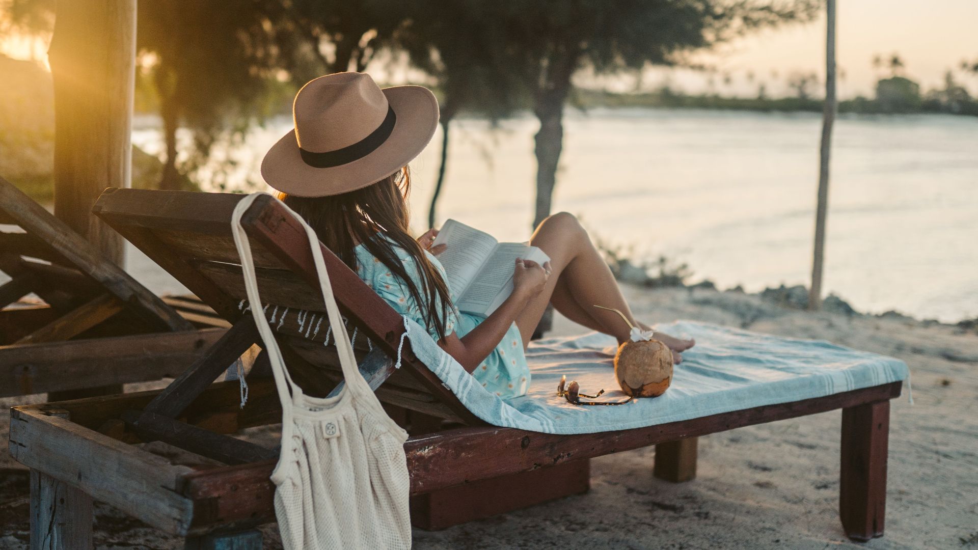 Woman reading on a beach lounger under a sun hat in Zanzibar at sunset, embracing vacation tranquility.
