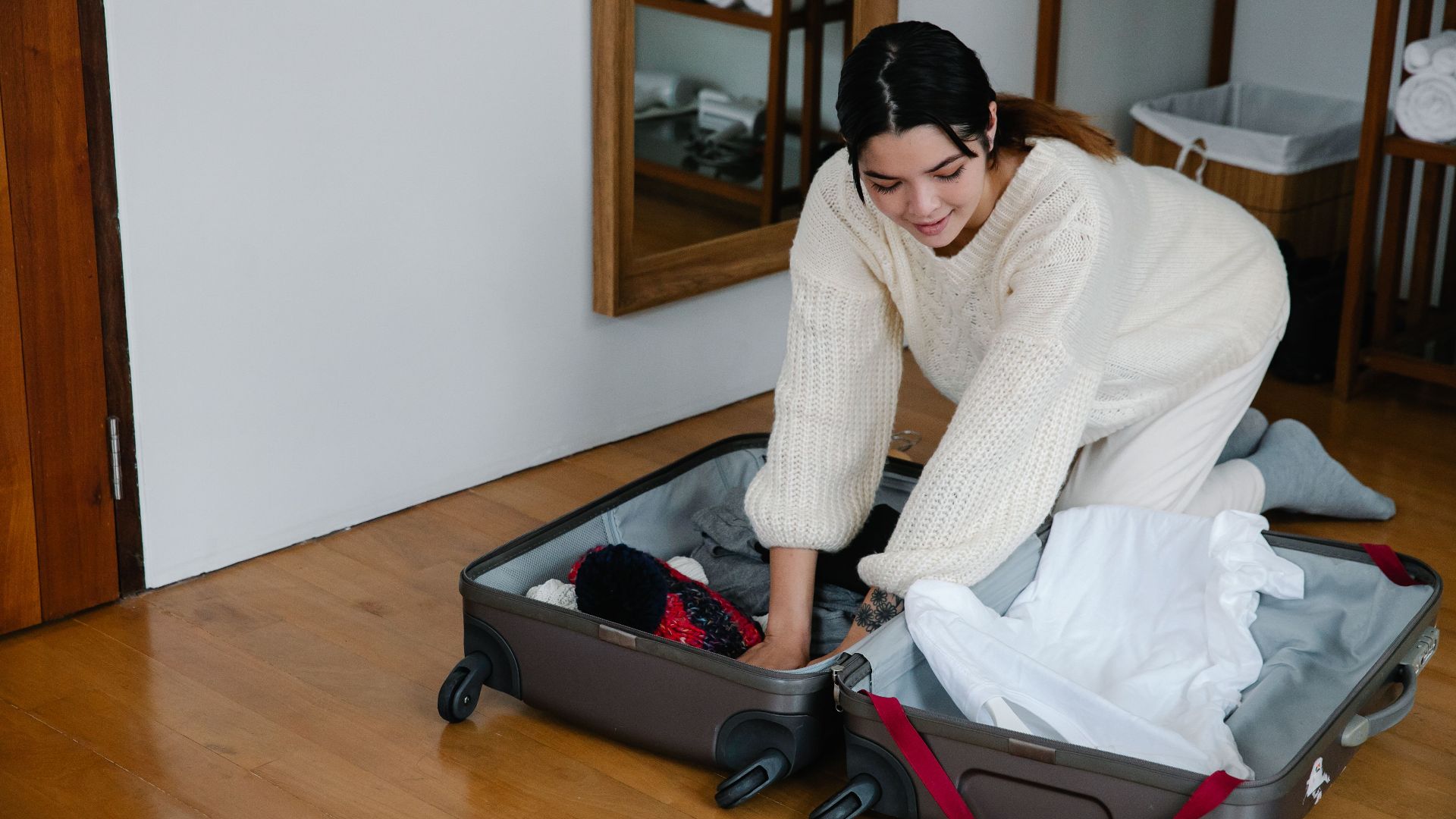 A woman kneels on the floor packing clothes into a suitcase in a cozy bedroom setting.