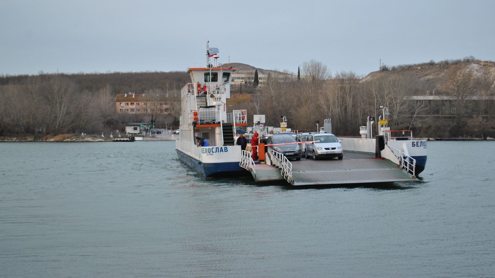 A ferry carrying cars across a calm river on a cloudy day, showcasing transportation in nature.