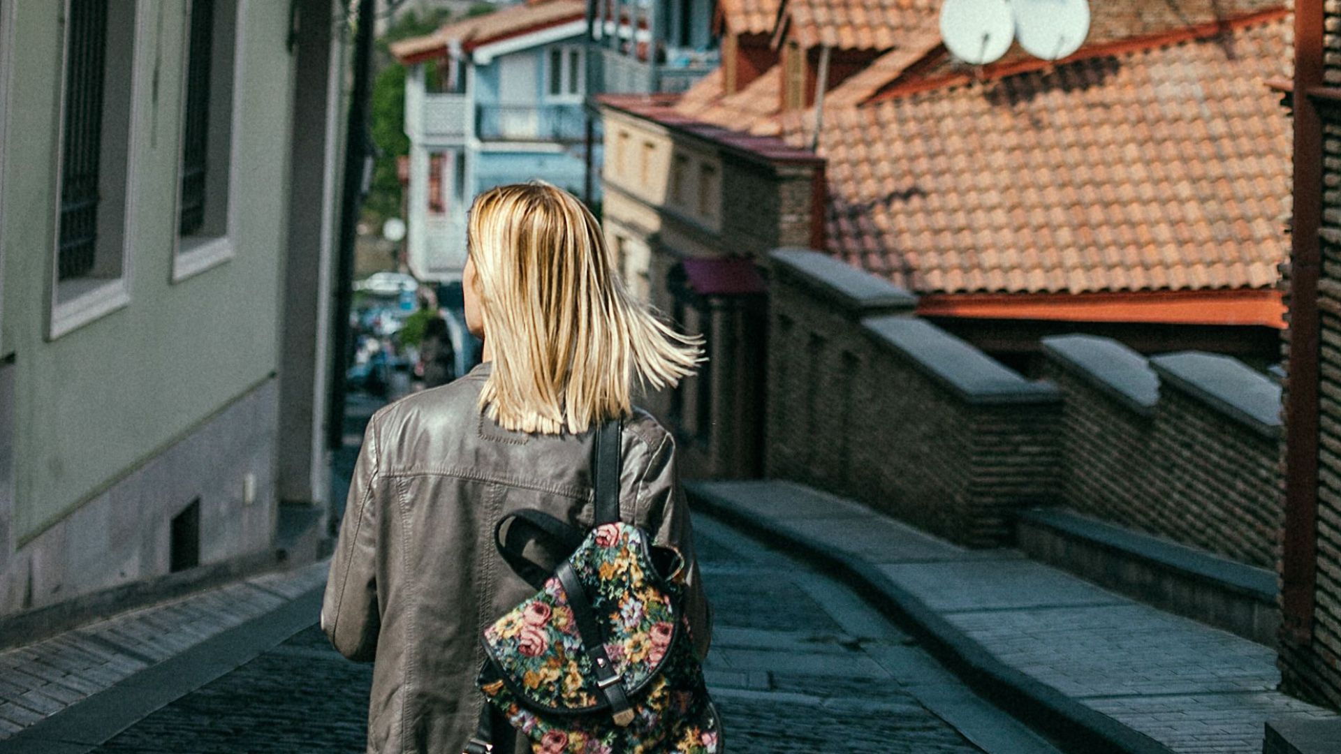 Back view unrecognizable female tourist with backpack walking on pedestrian cobblestone street between residential buildings in small town on sunny summer day