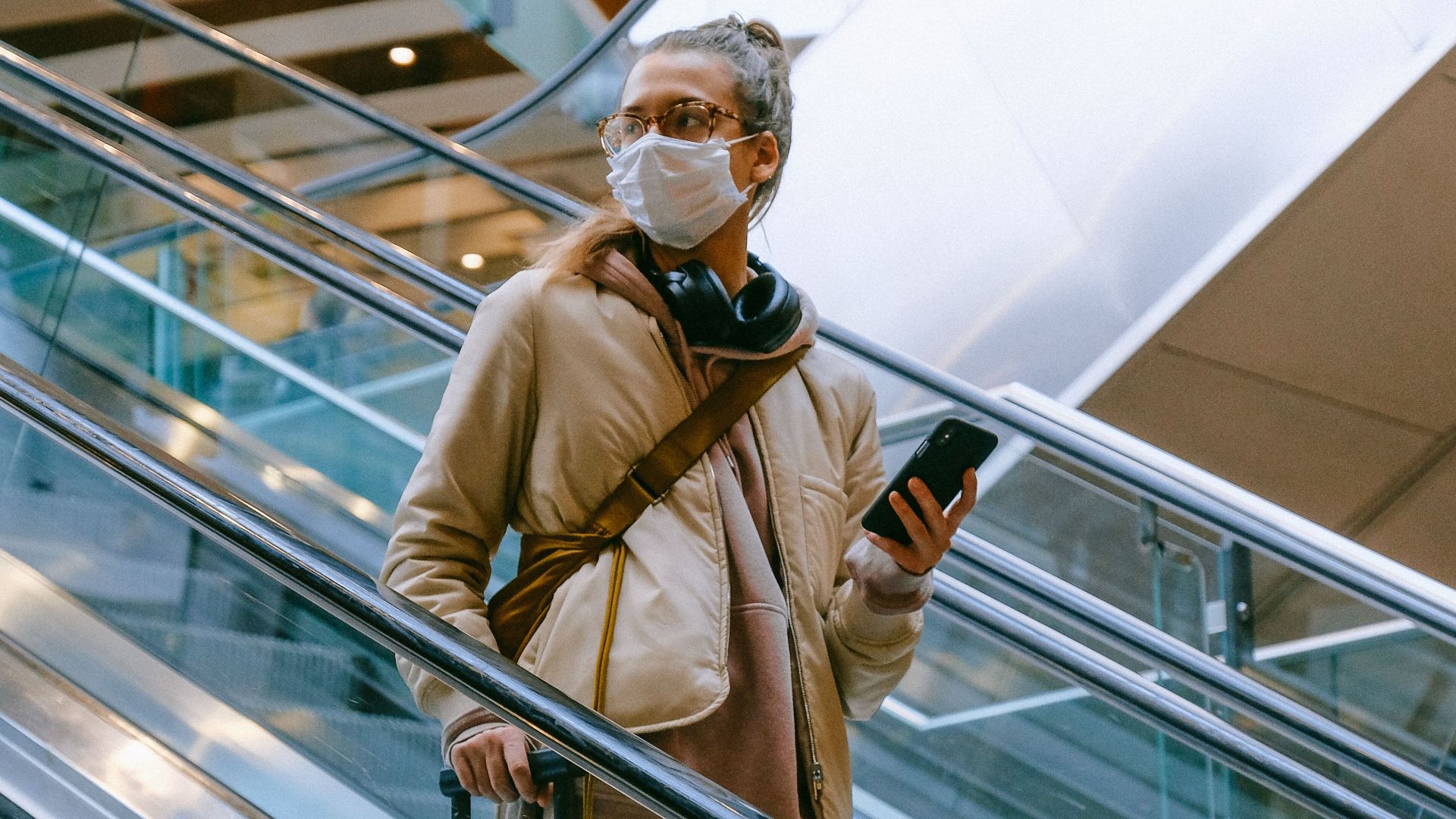 A woman wearing a mask uses an escalator in an airport with her luggage, reflecting modern travel norms.