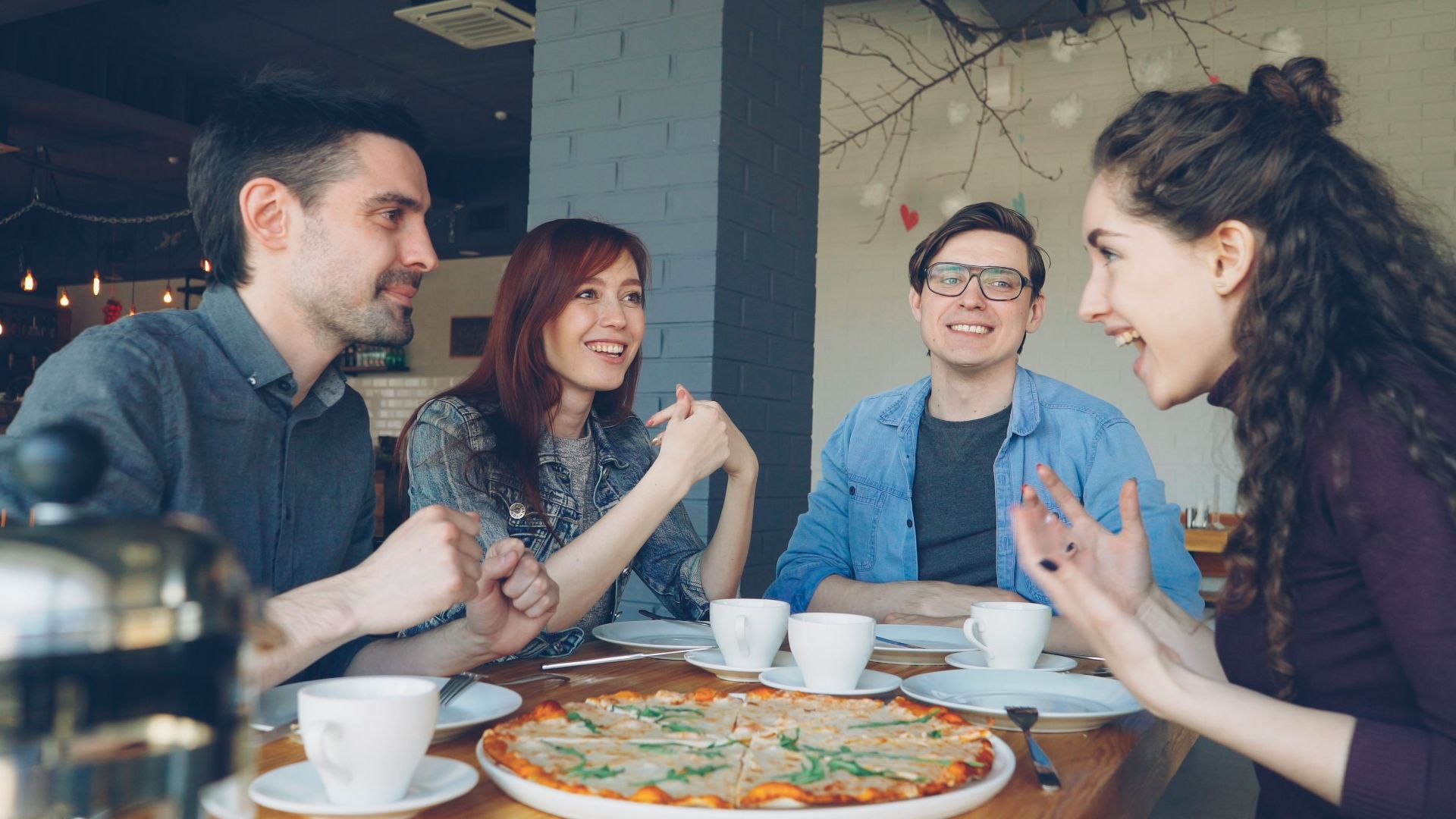 Group of four friends laughing and enjoying pizza and coffee together in a cozy cafe.