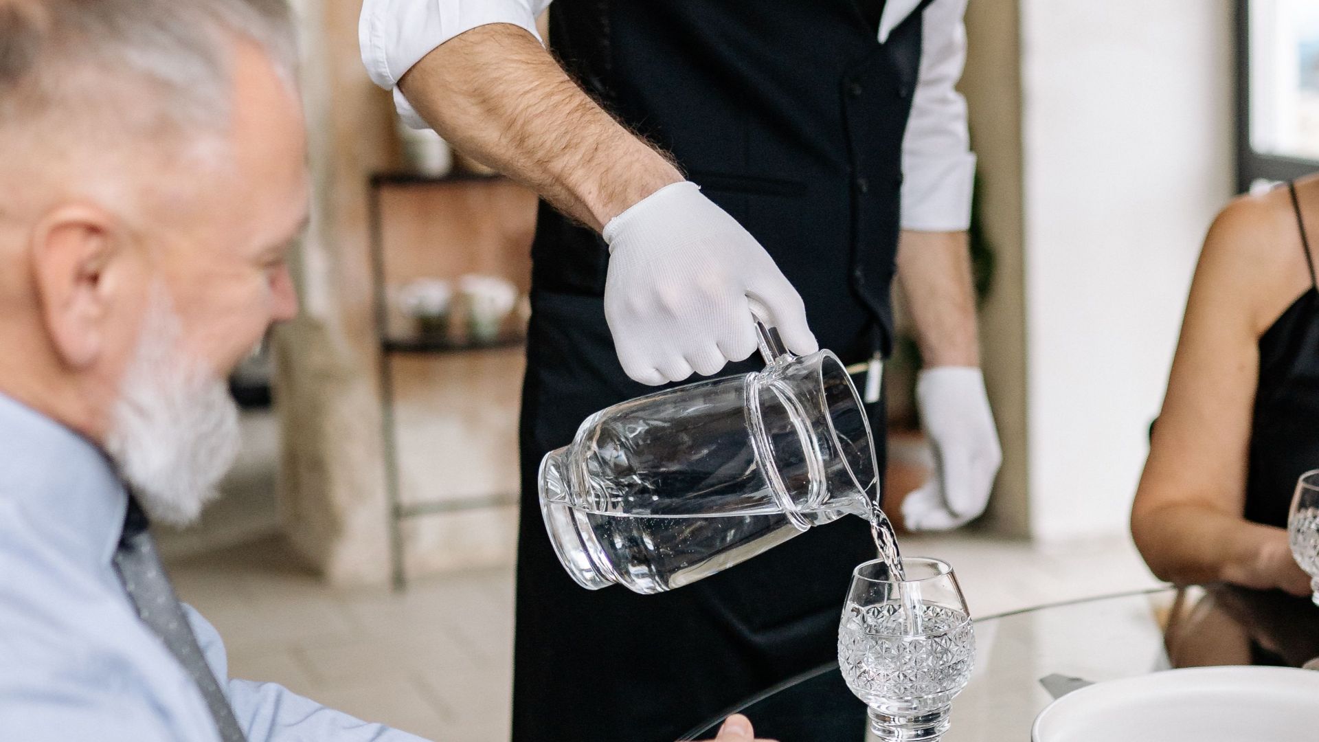A waiter wearing a mask serves water to an elderly couple in a restaurant setting.