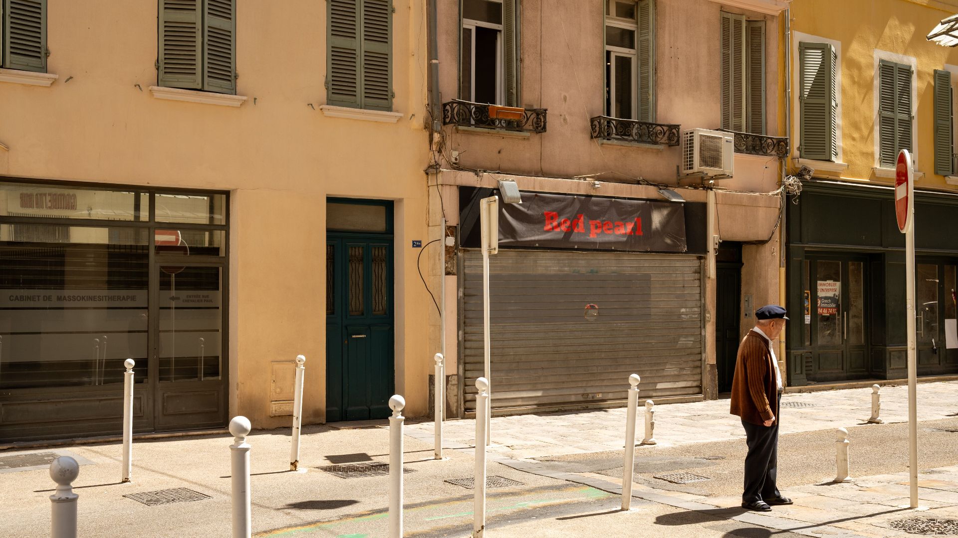 An elderly man walks past shuttered buildings on a sunny street in Toulon, France, capturing the calm urban scene.