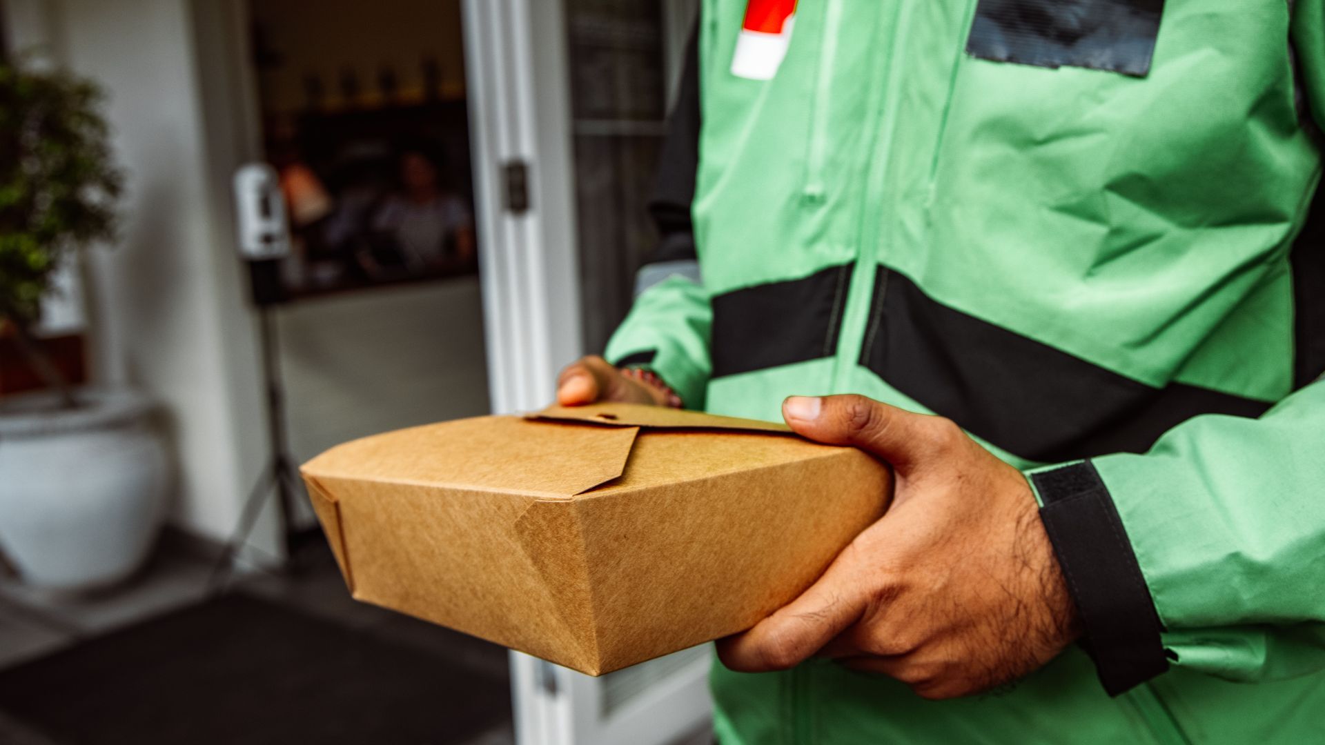 A deliveryman in a green jacket holding an eco-friendly food package outside a restaurant.