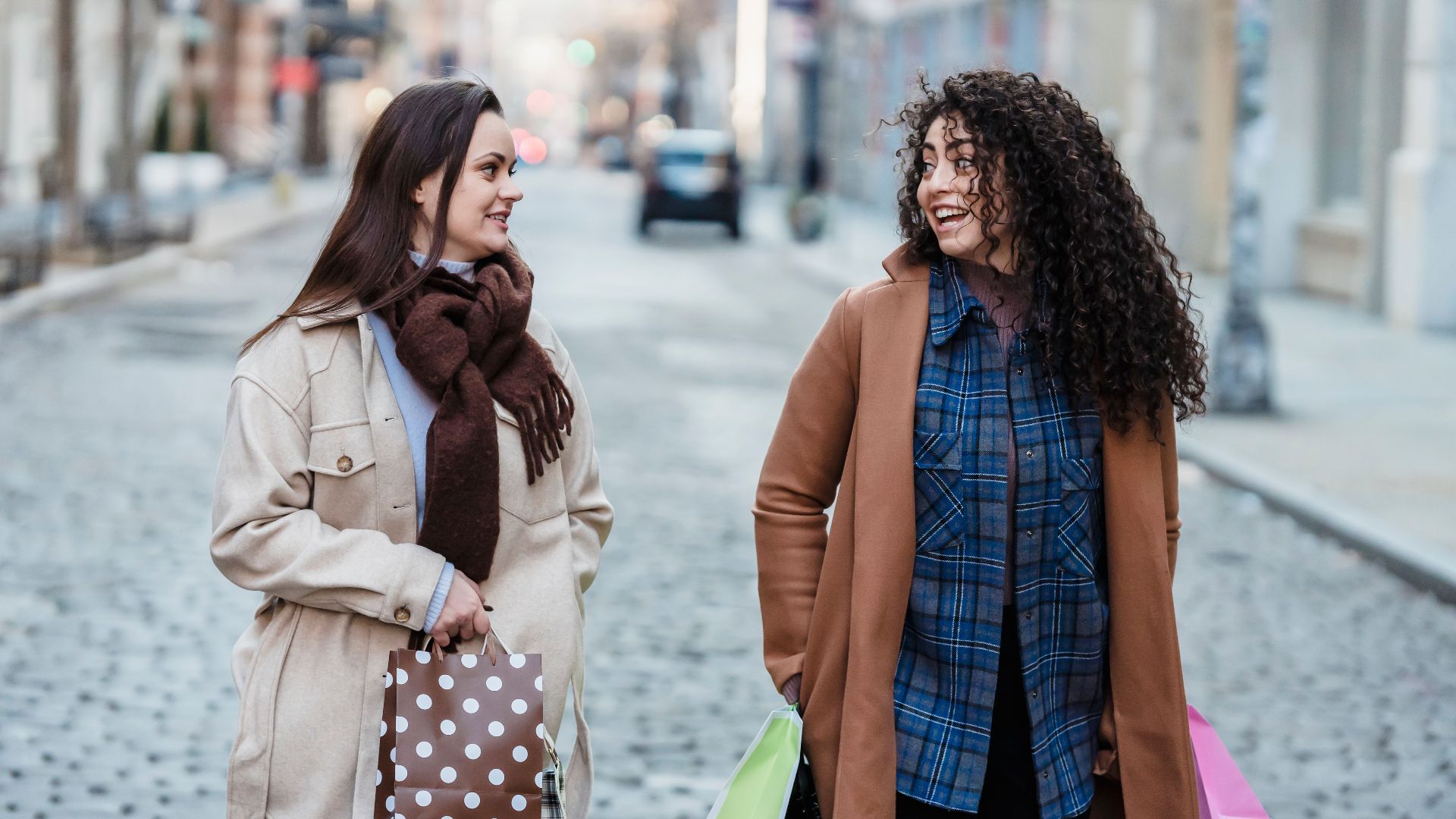 Positive multiracial female friends in elegant wear with gift bags looking at each other while strolling on sidewalk in city on blurred background