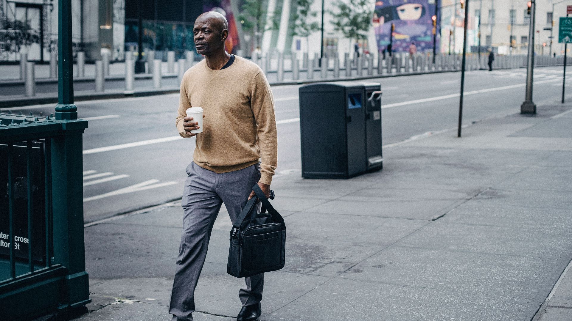 Full body of pensive African American male walking on pavement near asphalt road with takeaway coffee and black handbag in hand
