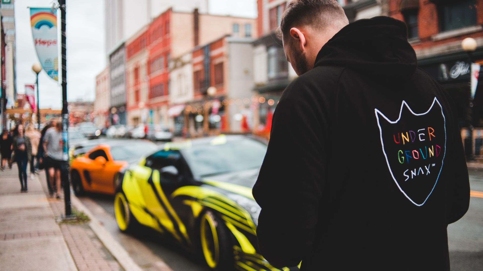 Back view of unrecognizable male in stylish hoodie with colorful inscription walking on street near road with parked sports cars in daytime