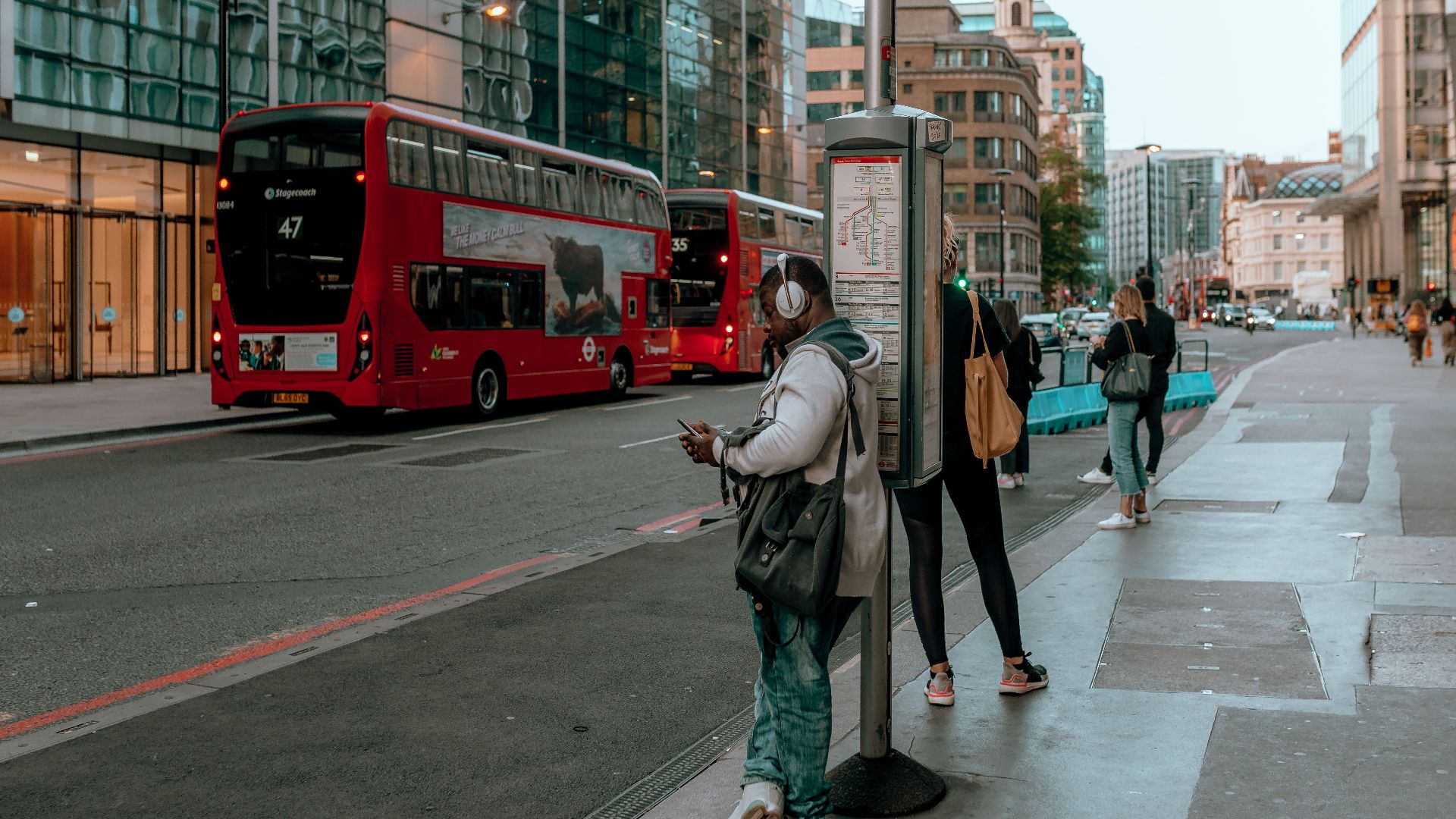 A busy street with people waiting at a bus stop and red double-decker buses passing by.