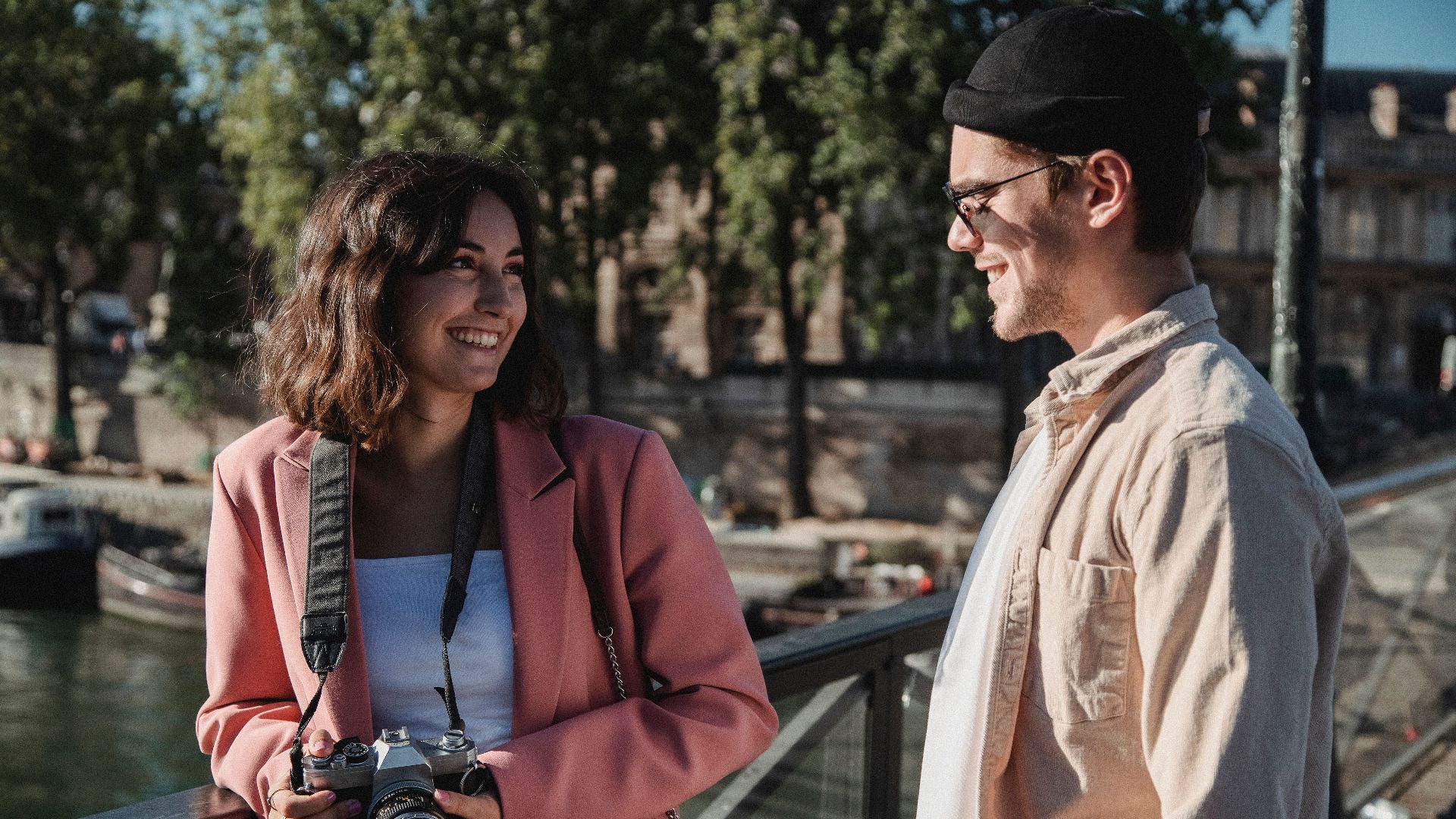 A happy couple smiling at each other with a camera in hand on a sunny day outdoors.