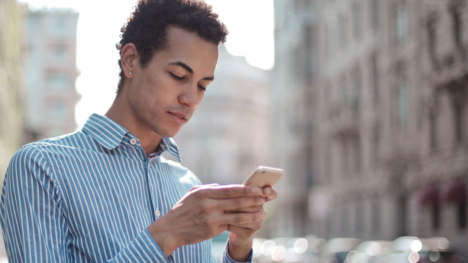 Focused young man in striped shirt browsing cellphone while standing in downtown on sunny day
