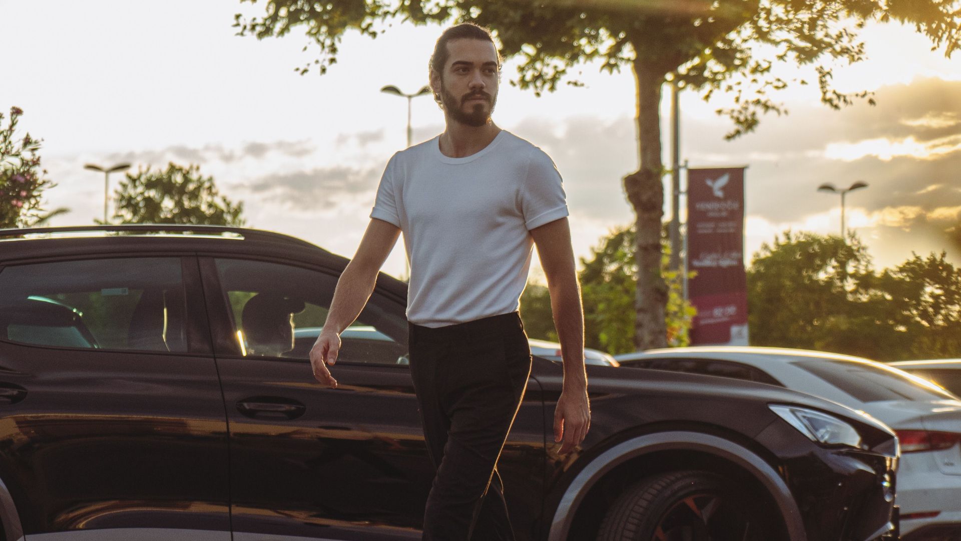 A man walks confidently past a black car outdoors in Istanbul, during golden hour.