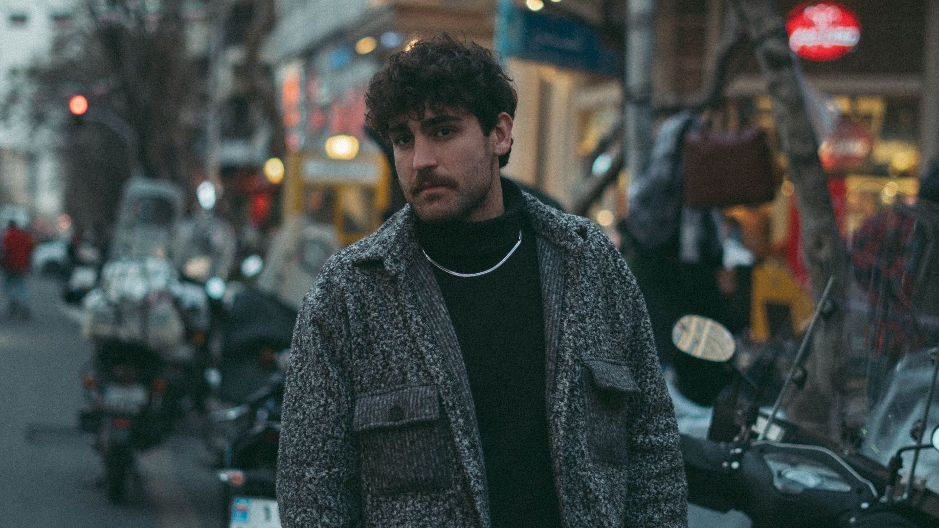 Stylish man in streetwear posing on a bustling city sidewalk during dusk.