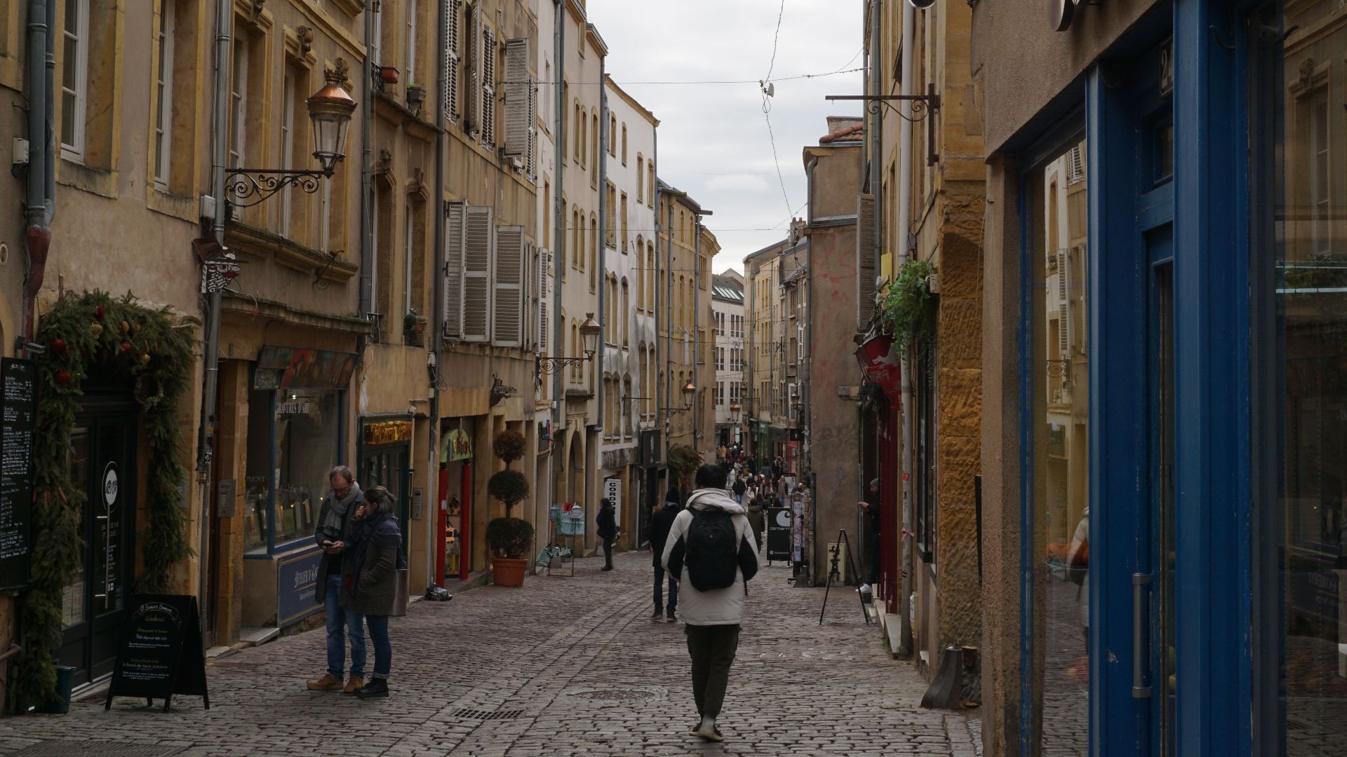 A quaint cobblestone street in an old European town with people walking under overcast skies.