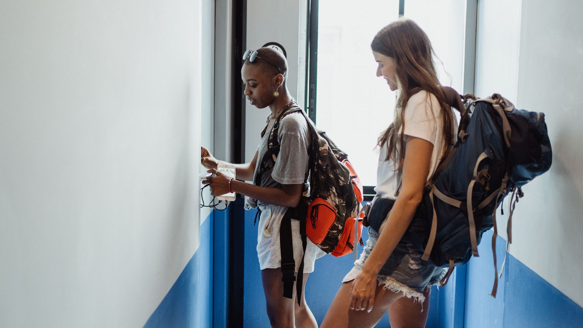 Two women travelers in a hostel corridor with backpacks, preparing for their journey.