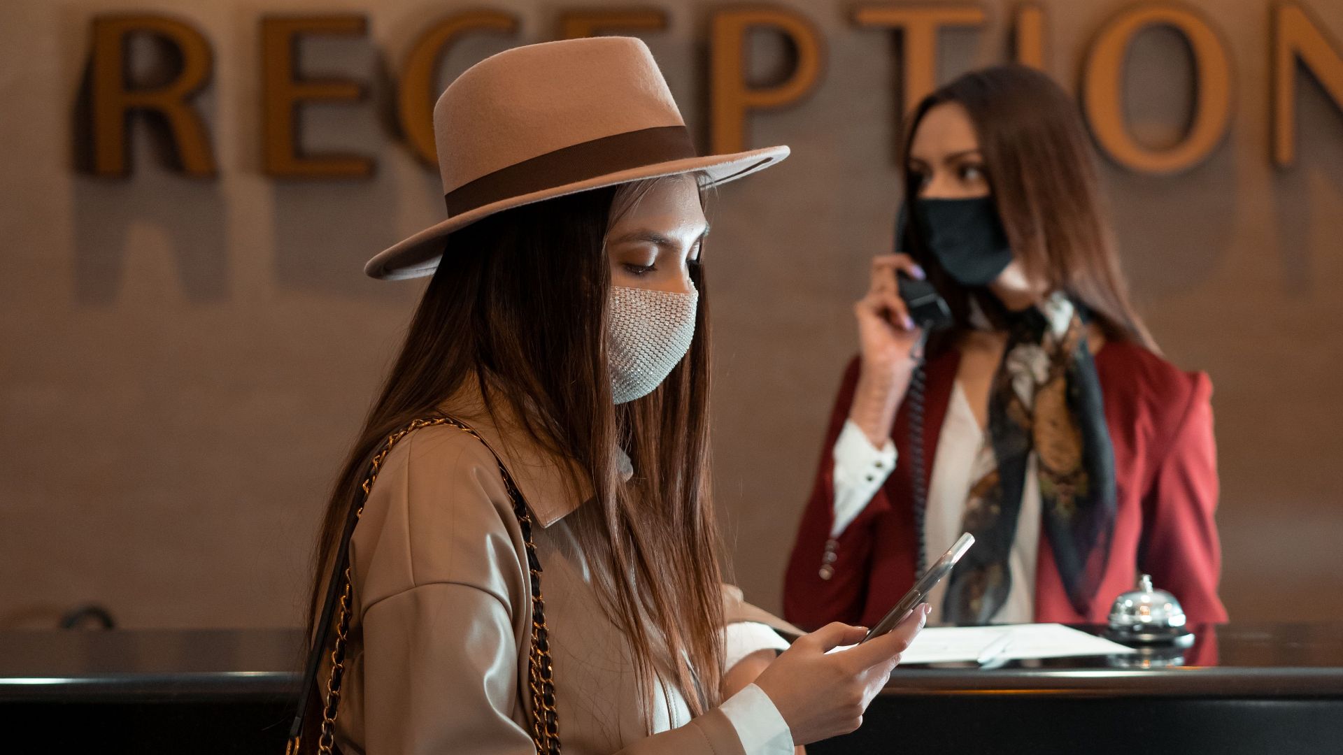 Woman in face mask at hotel reception counter checking her phone, symbolizing the new normal.