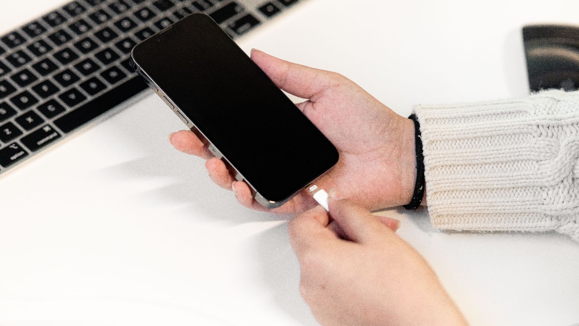 A woman plugs her smartphone into a charger at a desk, next to a laptop, illustrating modern technology usage.