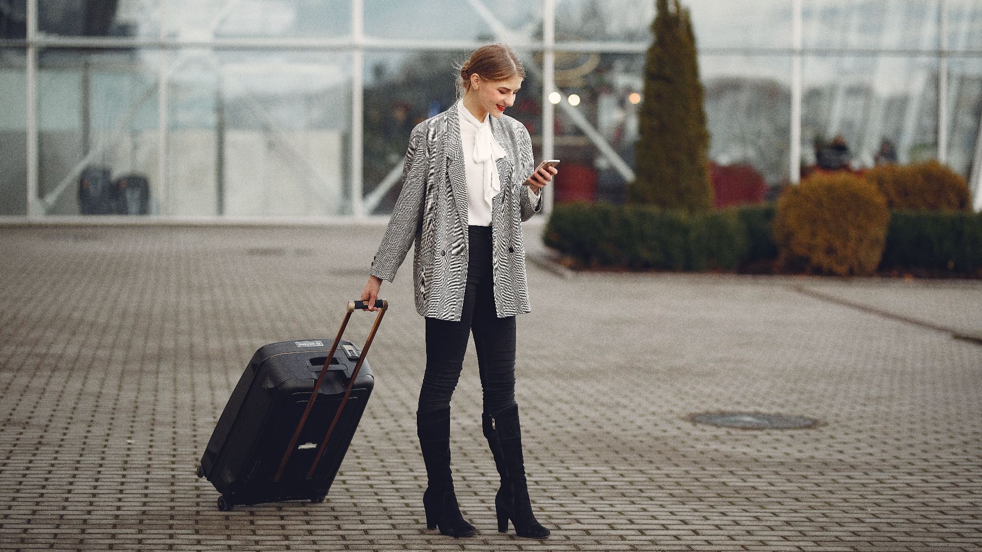 Full length of stylish woman in elegant outfit carrying baggage and surfing smartphone while standing near glass wall of airport