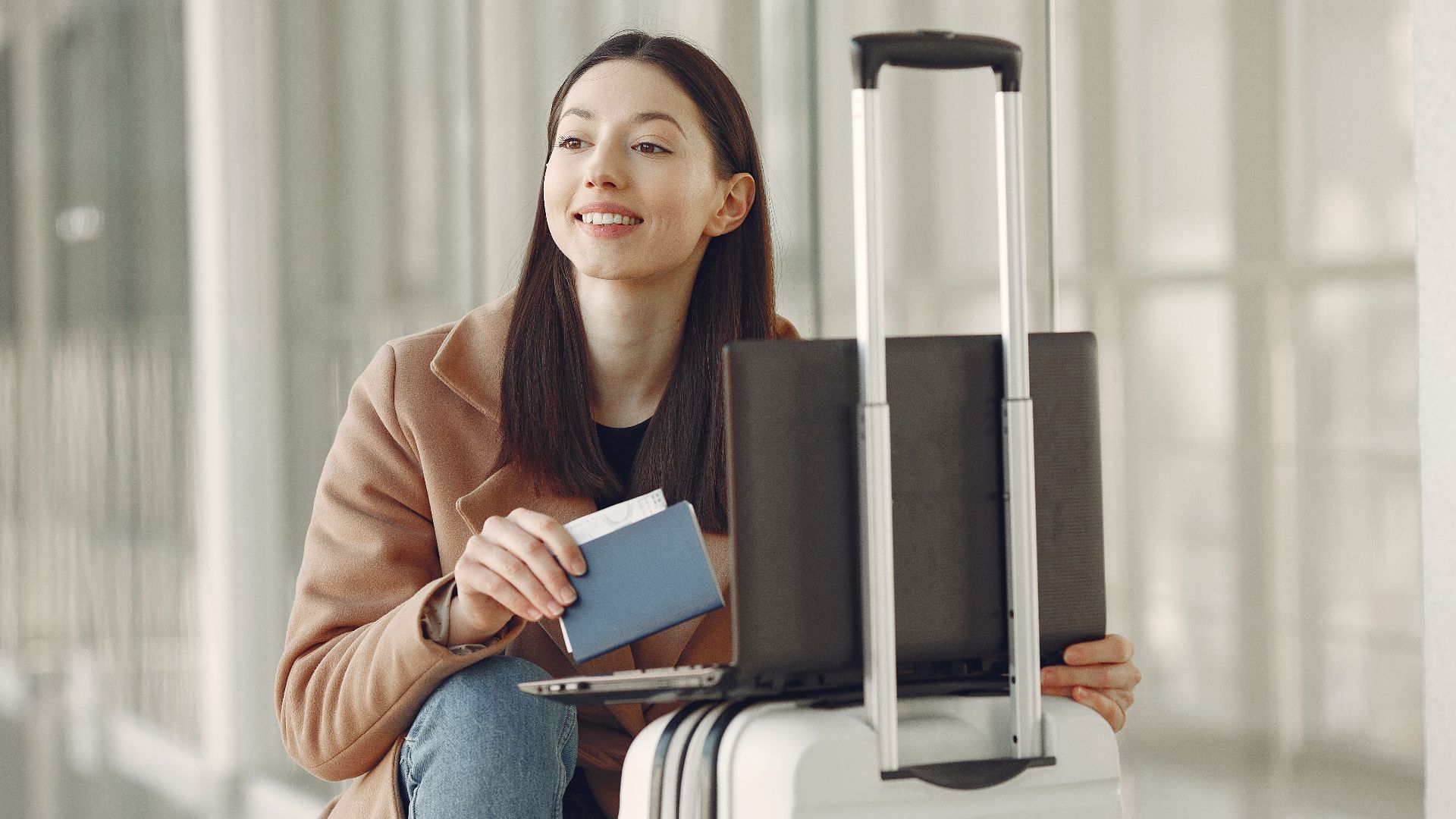Smiling adult ethnic female traveler in trendy coat holding passport and using laptop on luggage in airport corridor for checking ticket for correctness